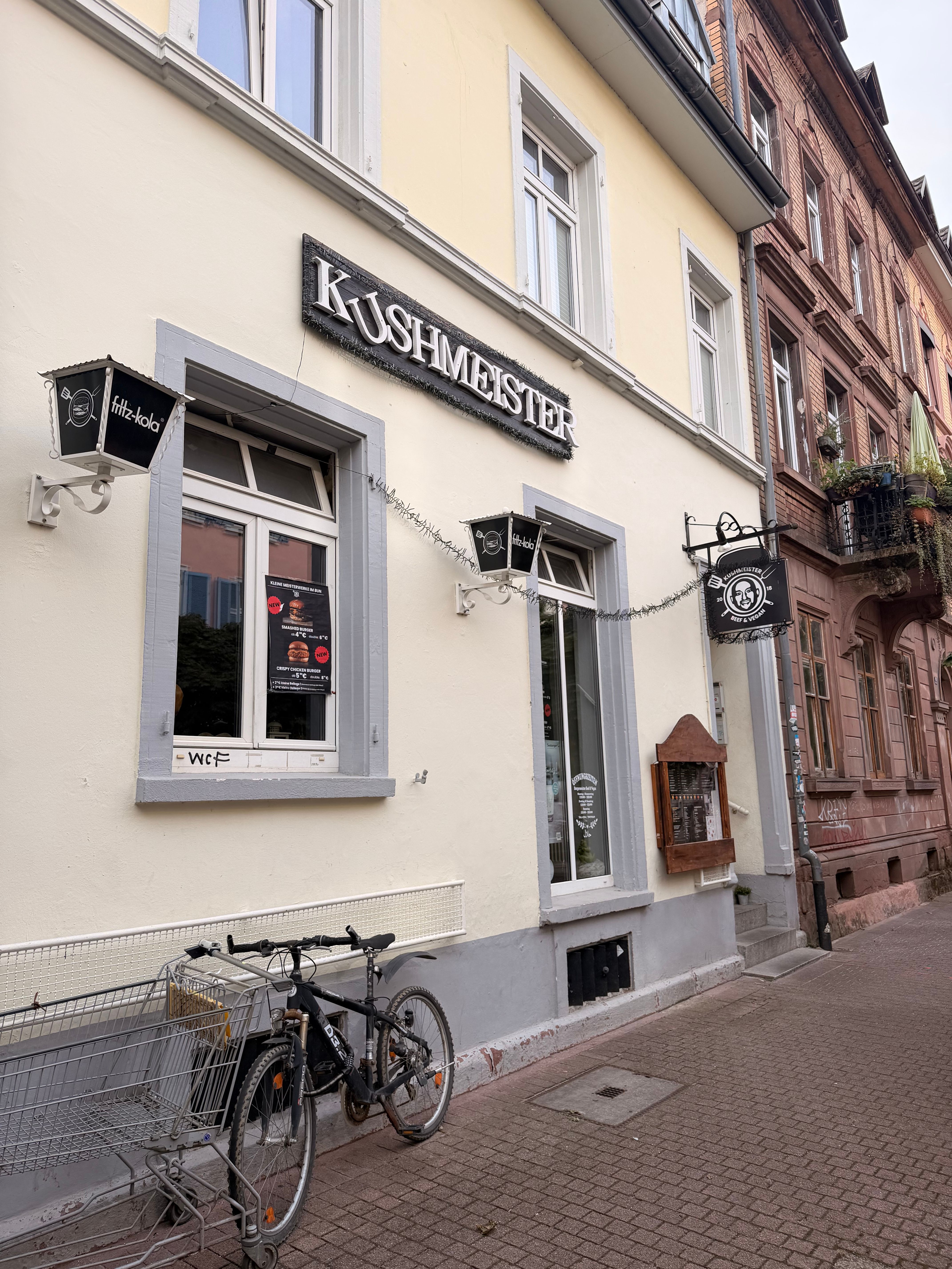 Fassade des Restaurants Kushmeister in einer schmalen Straße mit Schildern, Fenstern und abgestellten Fahrrädern vor dem Eingang.