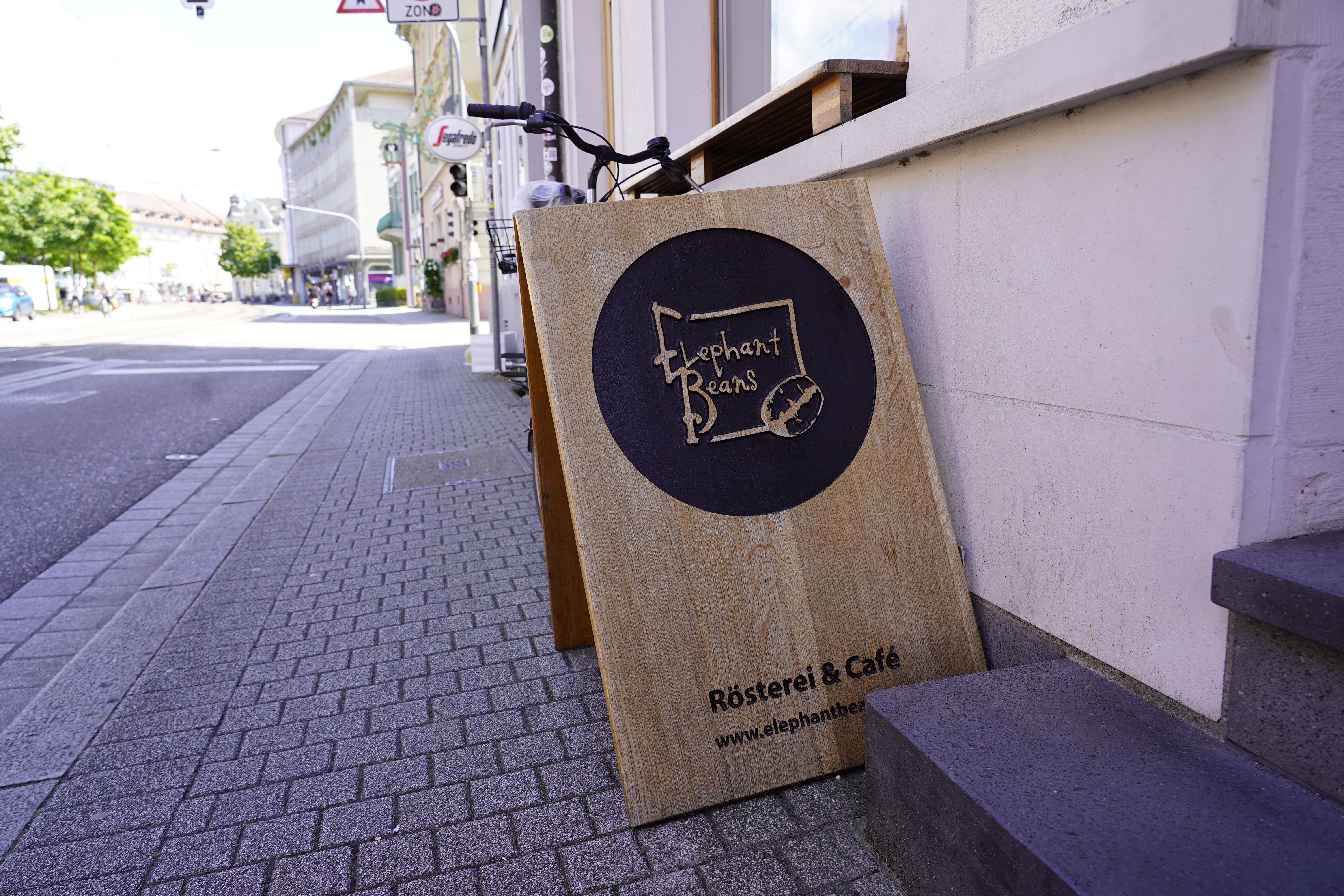 Holzschild mit 'Elephant Beans Rösterei & Café' an einer Hauswand in einer Stadtstraße.