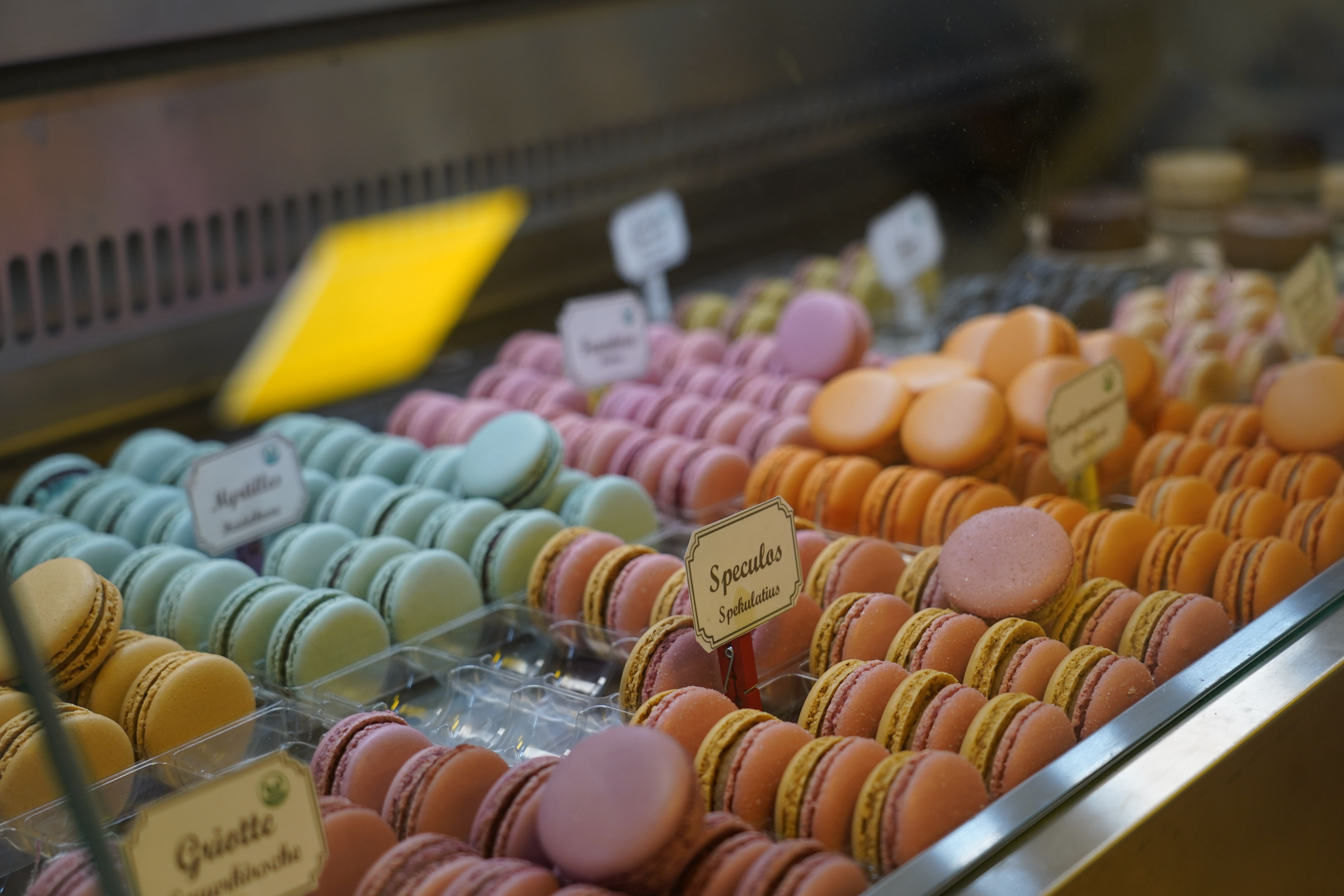 Bunte Macarons in einer Vitrine, sortiert nach Geschmacksrichtungen wie Spekulatius und Heidelbeere, in einer Patisserie ausgestellt.