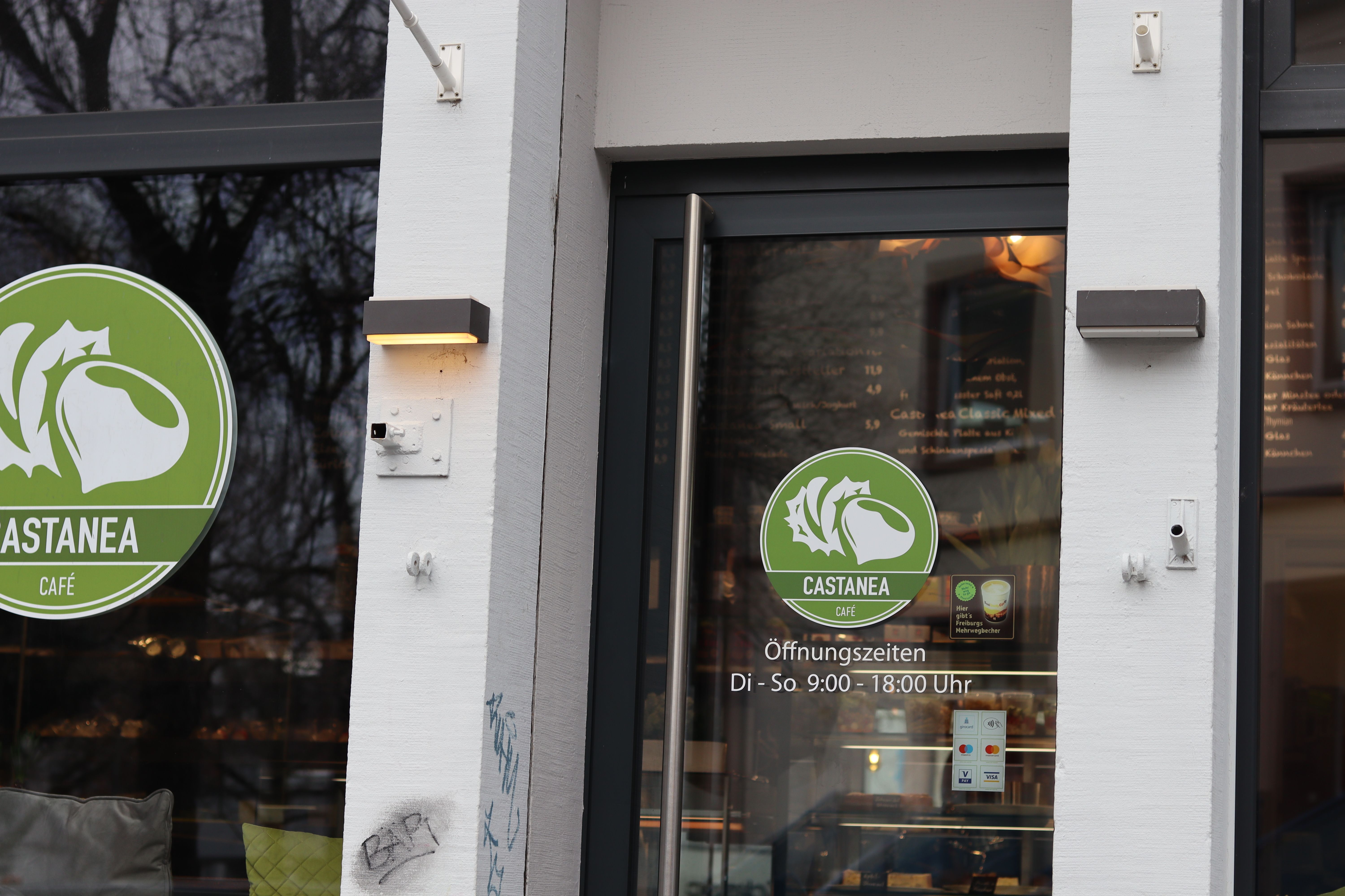 Eingang des Castanea Cafés in Freiburg mit grünem Logo und Öffnungszeiten. Die Tür ist aus Glas, daneben eine weiße Wand mit Beleuchtung.