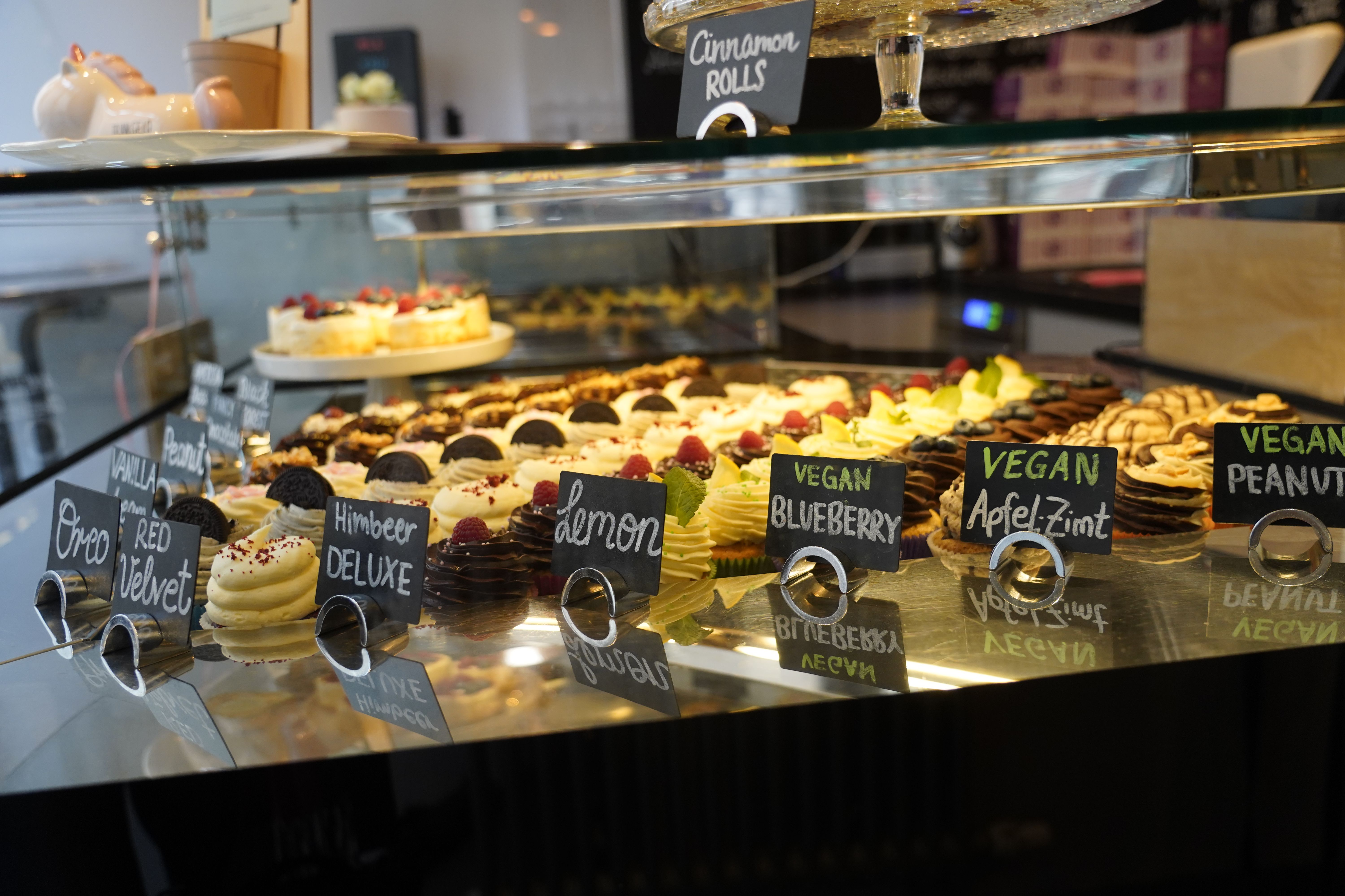 Vitrine mit Cupcakes in verschiedenen Geschmacksrichtungen wie Oreo, Red Velvet und veganen Sorten. Schilder zeigen die Namen der Geschmacksrichtungen an.