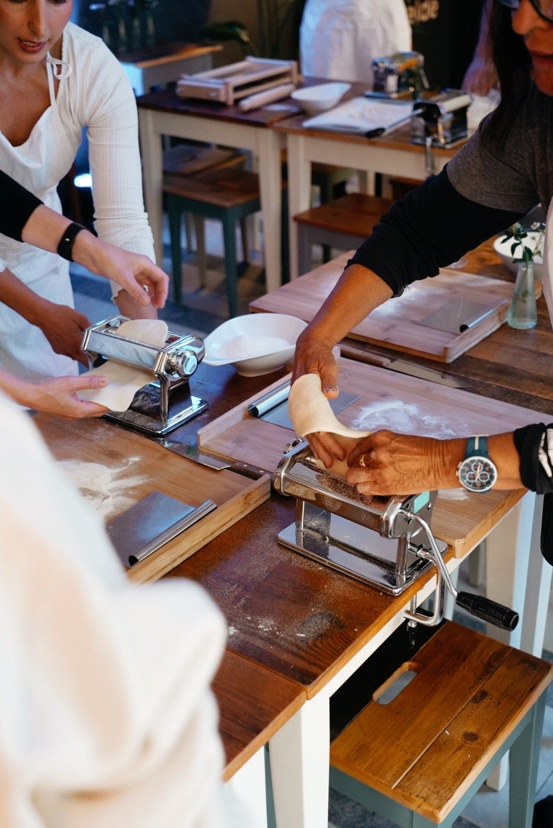 Mehrere Personen bereiten an Holztischen frische Pasta mit Nudelmaschinen zu. Der Raum wirkt wie eine Kochschule oder ein Workshop.