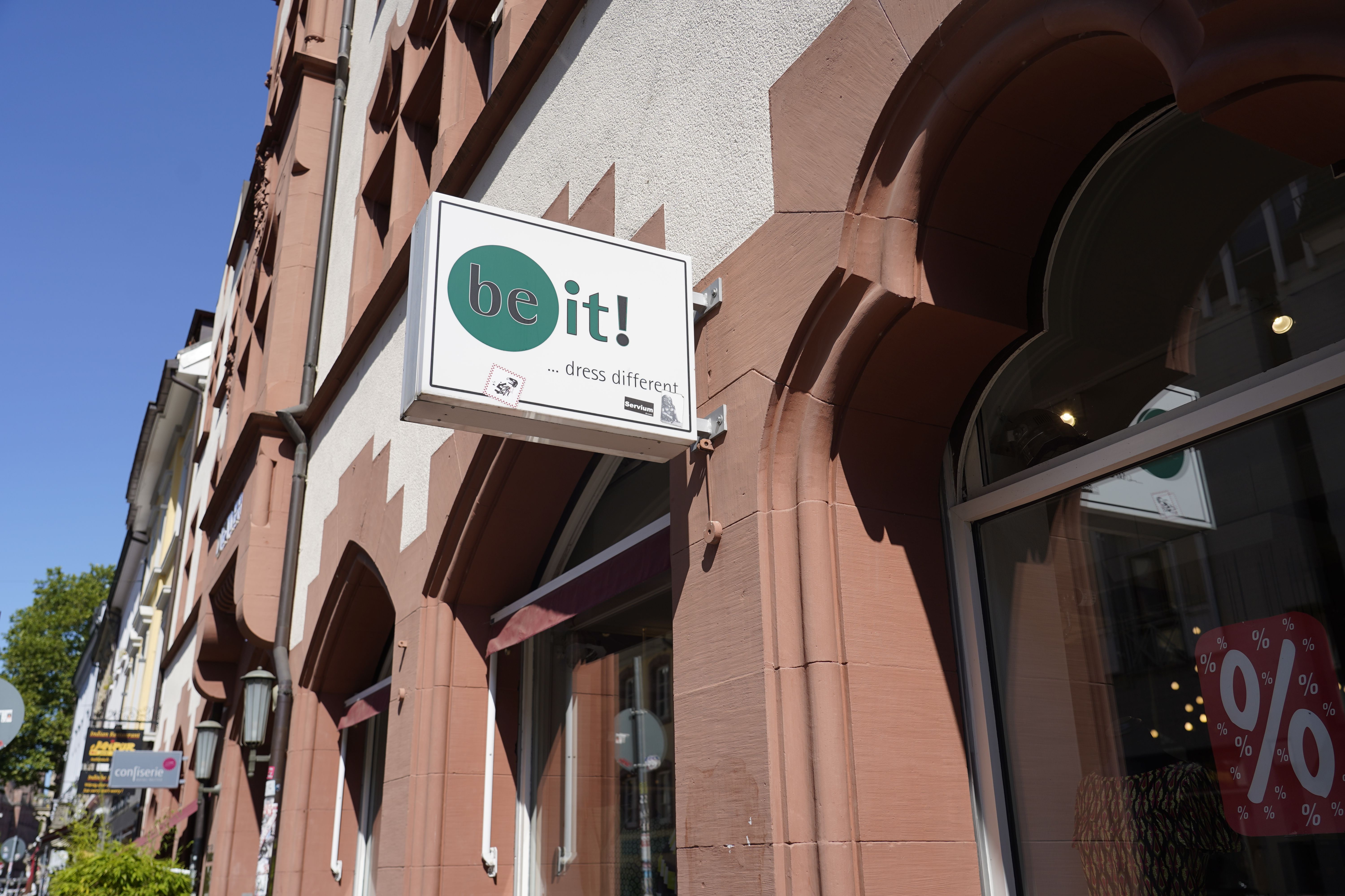Geschäftsschild 'be it! ... dress different' an einer historischen Hausfassade bei sonnigem Wetter.