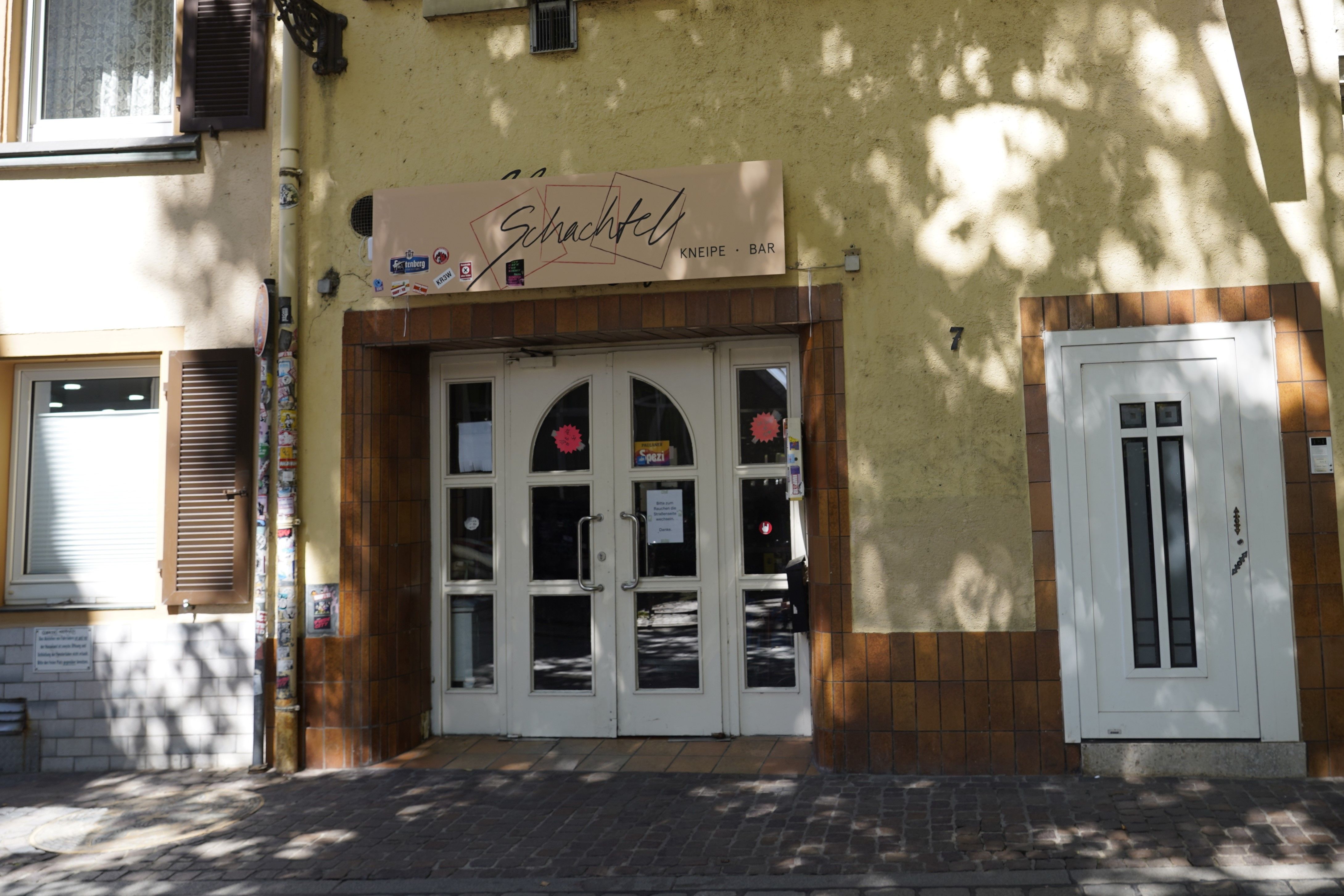Eingang der Kneipe Schachtel in Freiburg mit gelber Fassade, braunen Fliesen und einem Schild über der Tür.