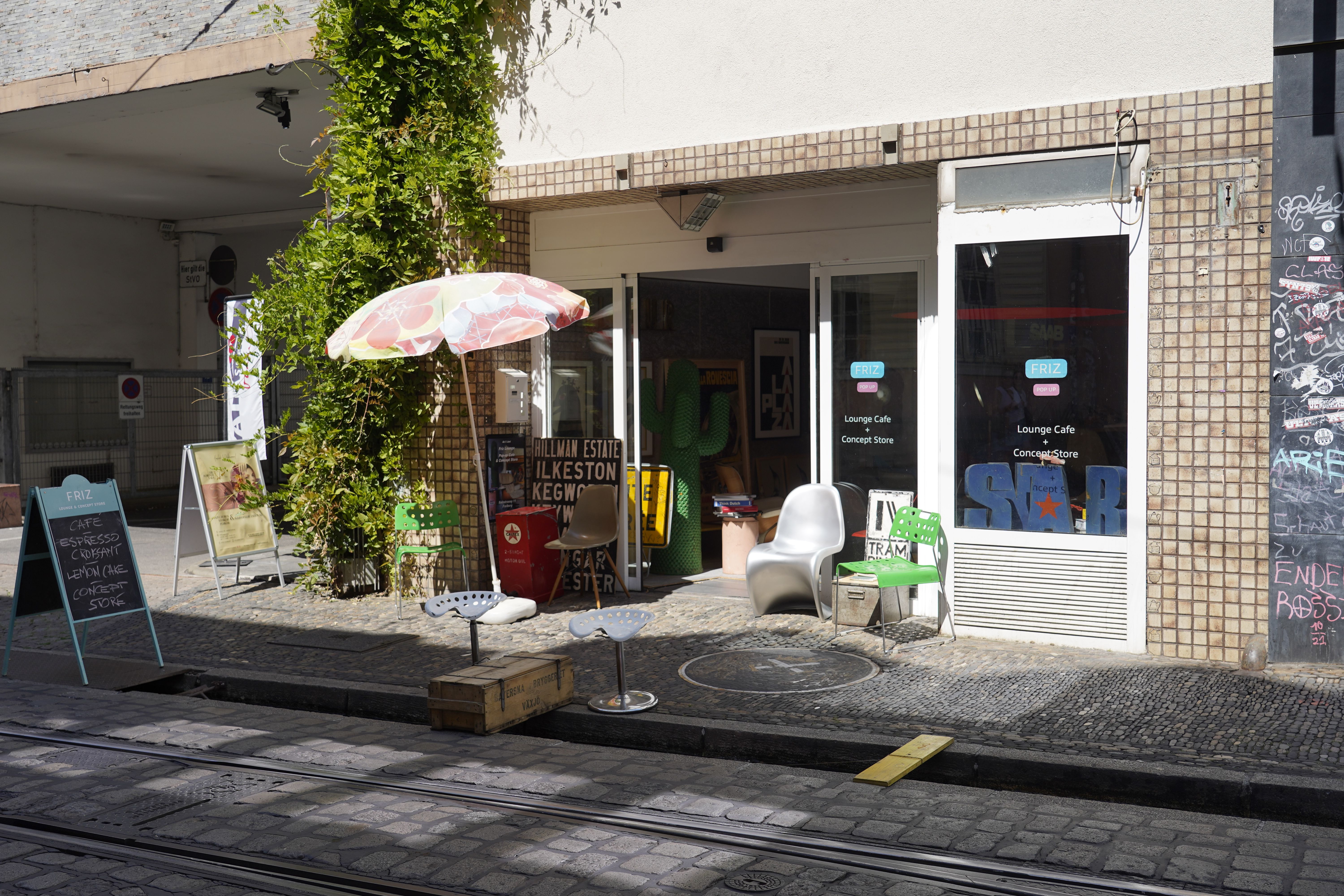 Eingang eines Concept Stores mit buntem Sonnenschirm, Stühlen und Schildern auf dem Gehweg. Die Fassade ist mit Kacheln verkleidet.