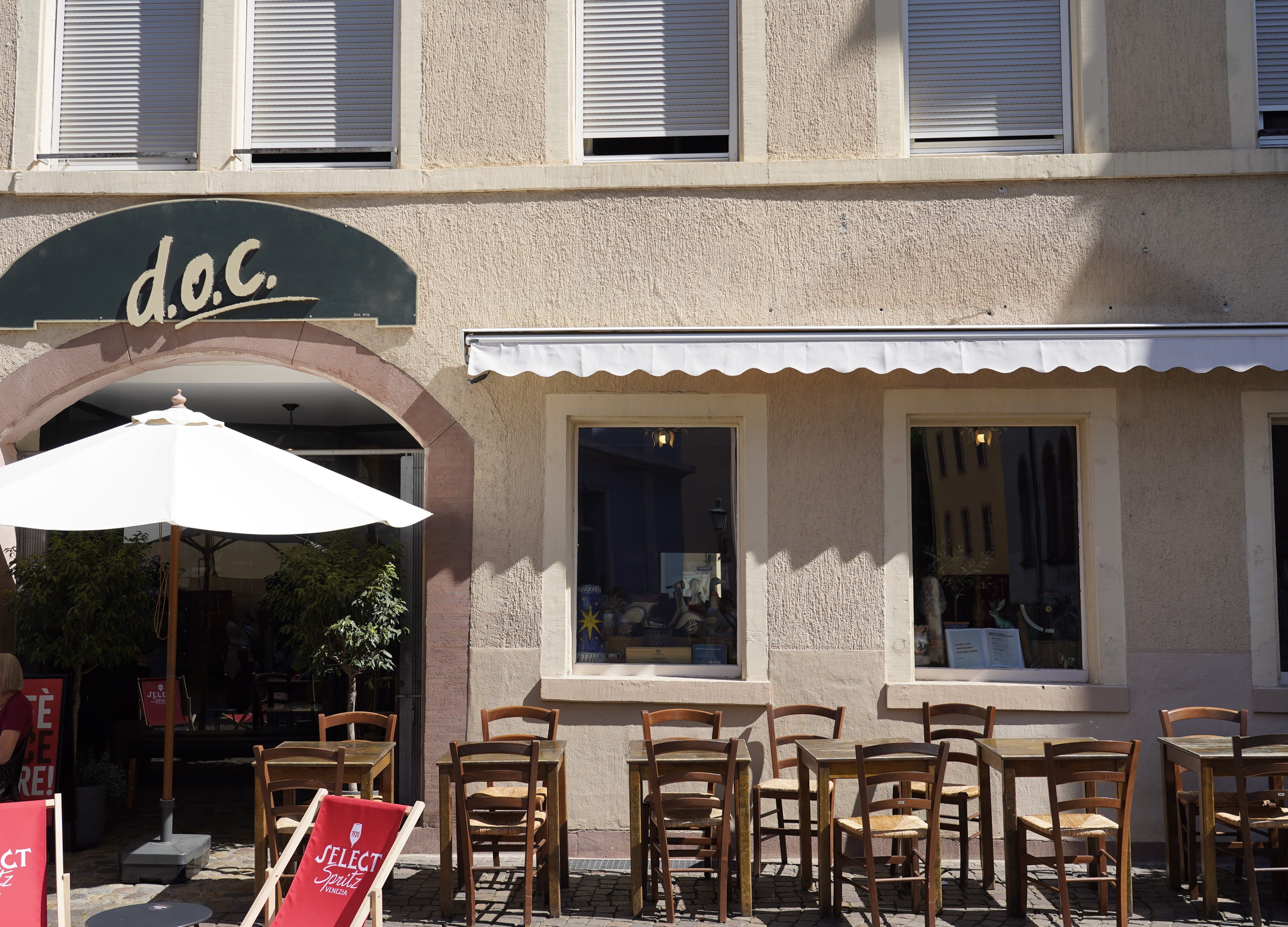 Außenansicht des Café mit Holztischen und Stühlen auf dem Gehweg. Ein Sonnenschirm spendet Schatten, und eine Markise ist ausgefahren.