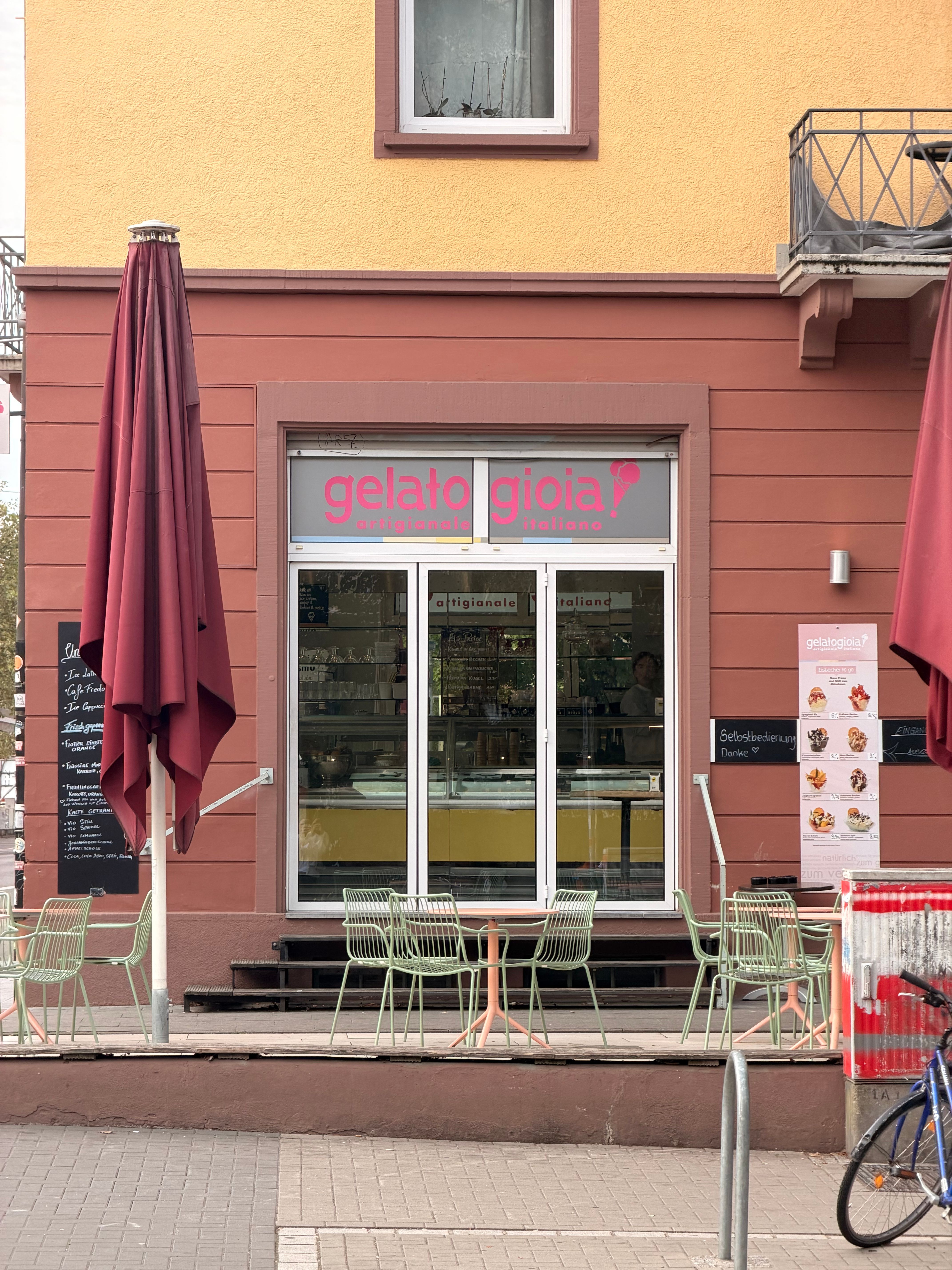 Eisdiele „gelatogioia“ mit rosa Schriftzug, Sonnenschirmen und Sitzplätzen vor einem zweifarbigen Gebäude.