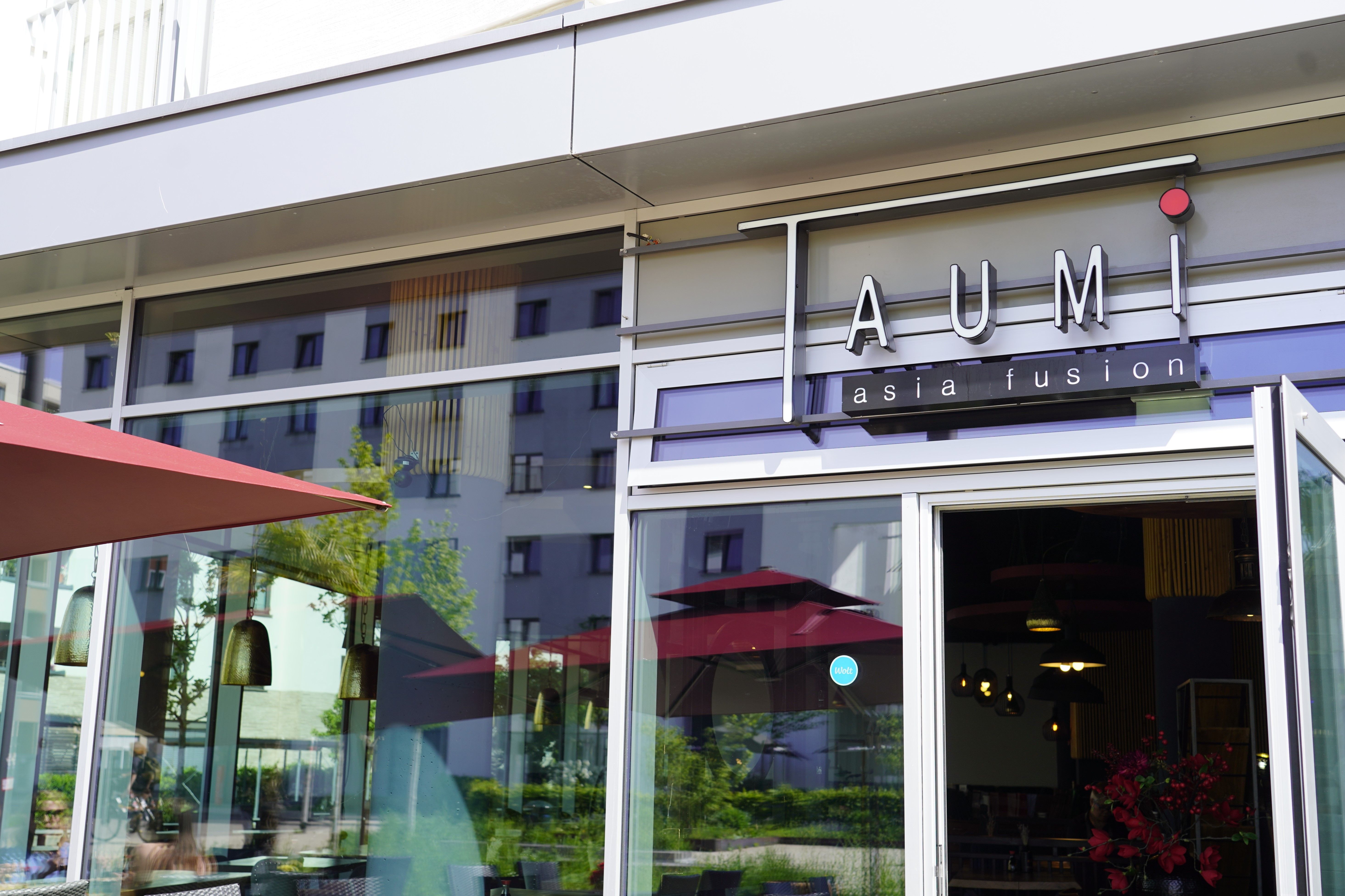 Eingang des Restaurants Taumi Asia Fusion in Freiburg. Geöffnete Türen, Spiegelung von Bäumen und Gebäuden im Fenster. Rote Sonnenschirme im Außenbereich.